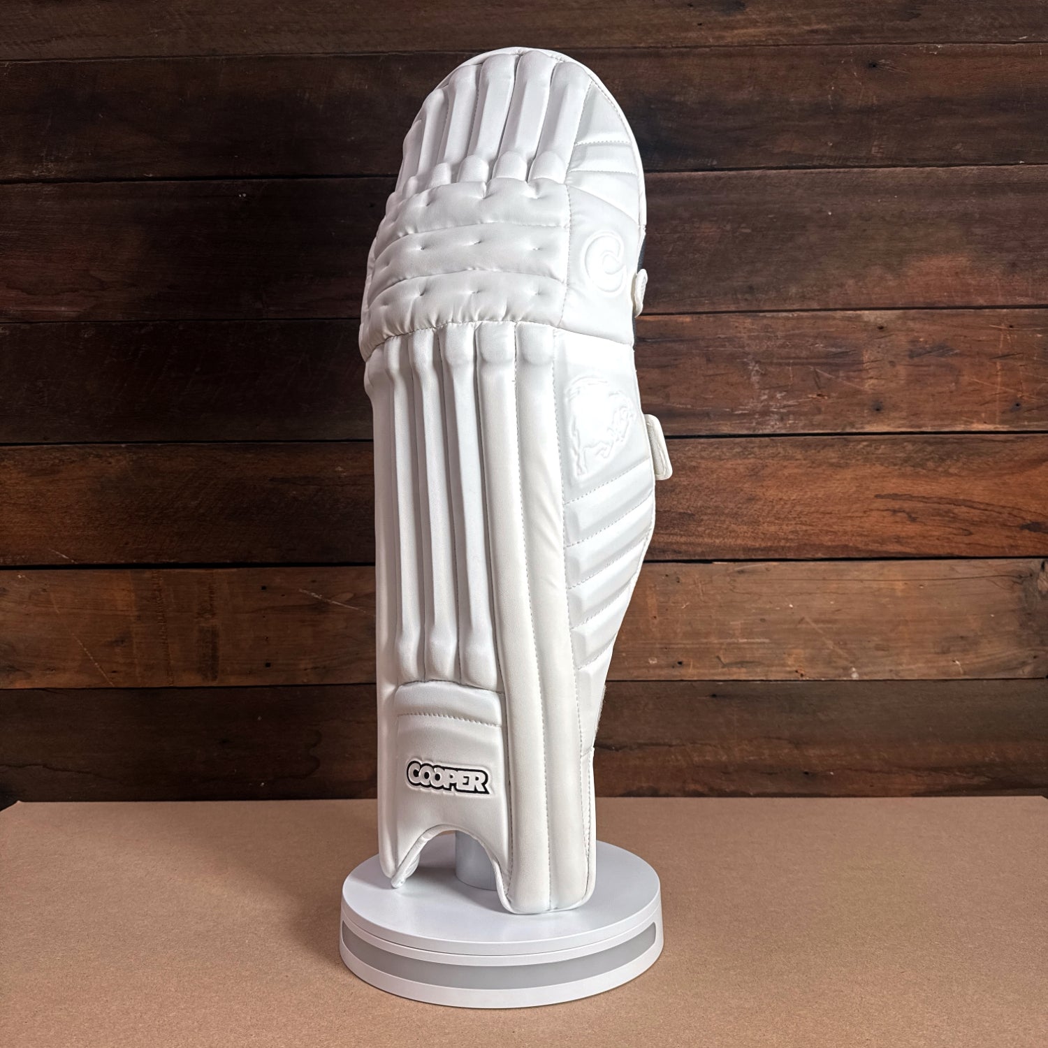 White cricket pad displayed on a stand against a wooden background, showcasing its protective design and branding.