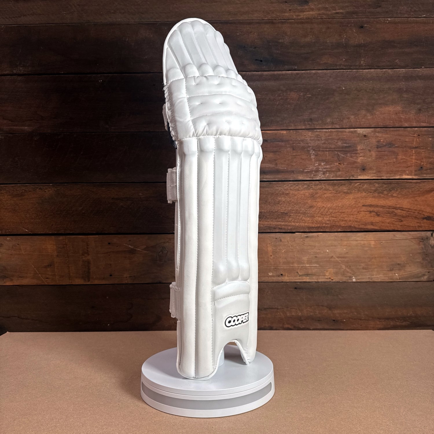 White cricket pad displayed on a stand against a wooden background, featuring protective design and branding.