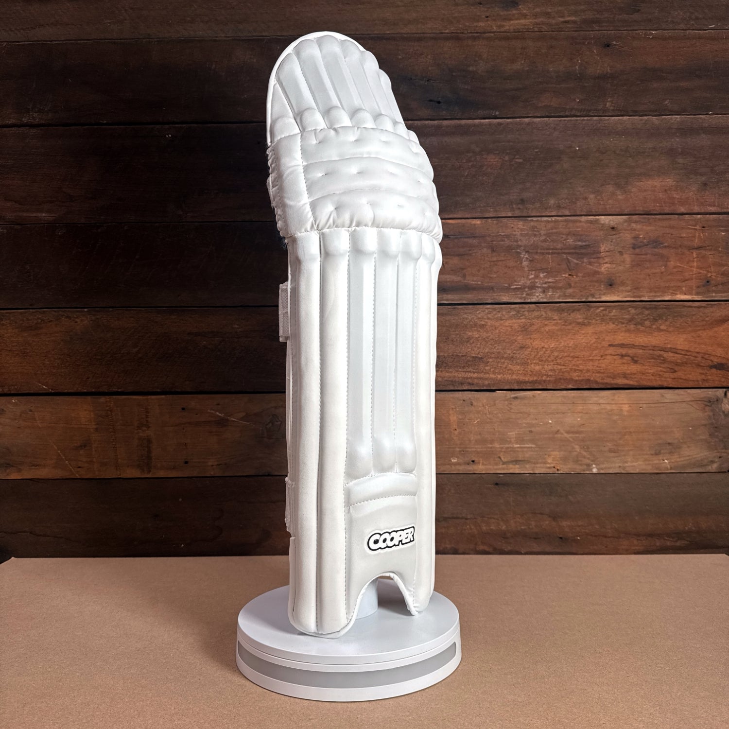 White cricket pad displayed on a stand, featuring Cooper branding and a sleek design on a wooden background.