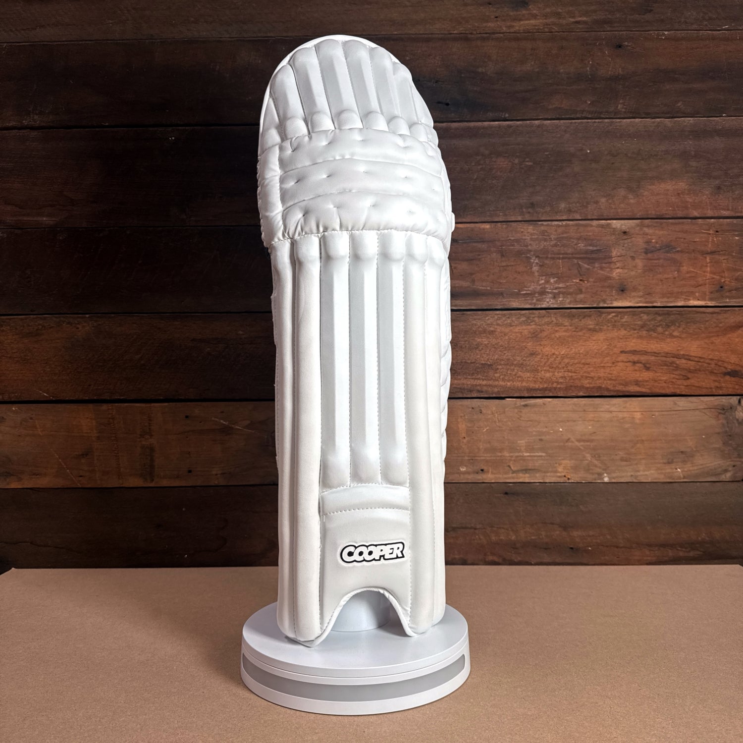 Cooper cricket pad displayed on a pedestal against a wooden background, showcasing premium design and protection.