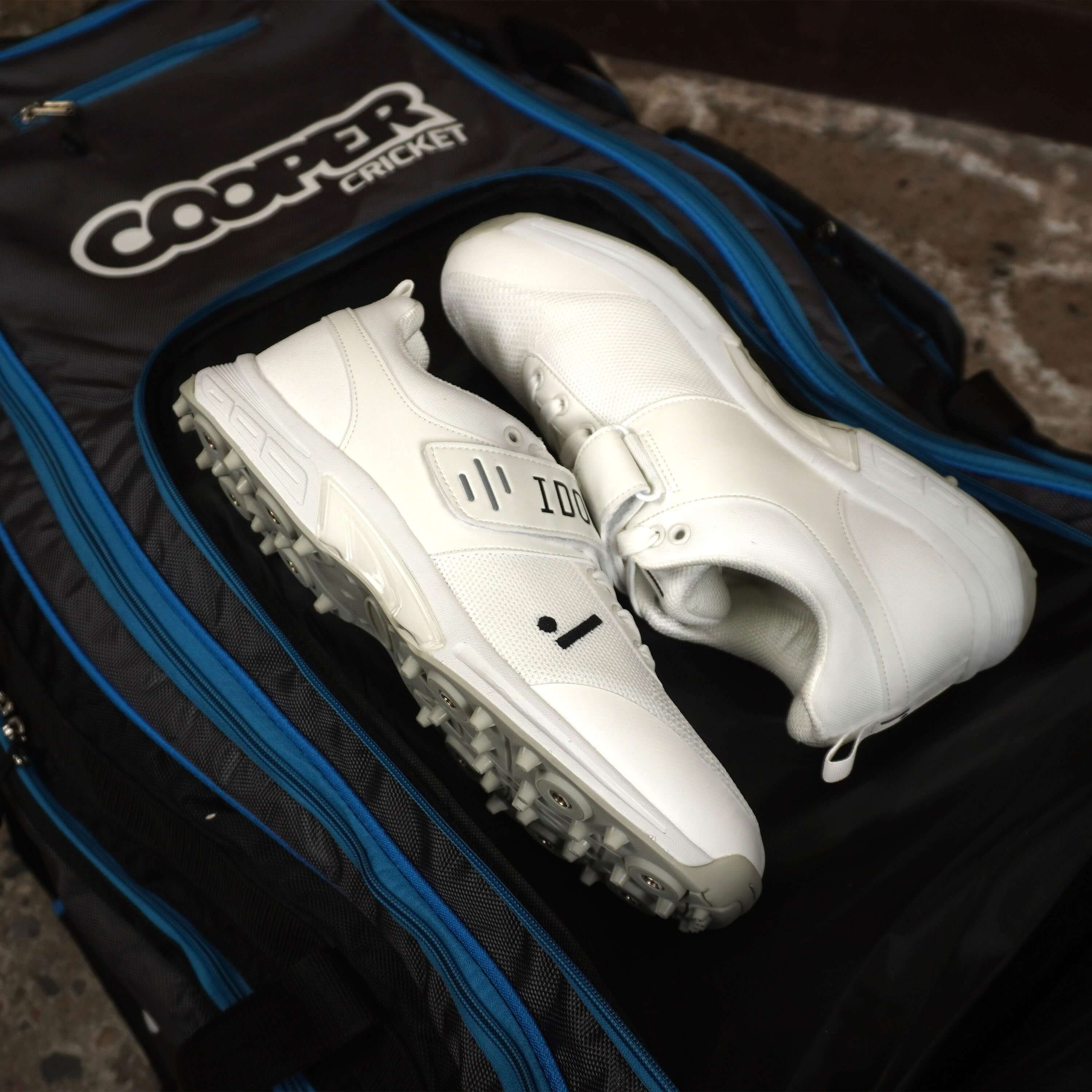 White cricket shoes resting on a black LK9 cricket bag with blue accents and multiple compartments.