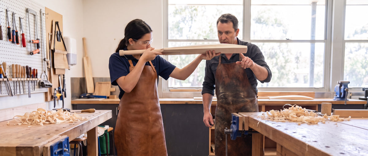 A teacher and student in leather aprons checking a freshly shaped cricket bat in a school workshop